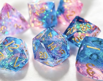 Puede incluir: Un juego de siete dados poliédricos que presentan una mezcla translúcida de tonos azules y rosas con escamas de papel iridiscente incrustadas. Cada dado tiene bordes afilados y números de color dorado, incluido un dado de veinte caras central.