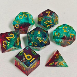May include: Set of seven polyhedral dice in various shapes. The dice are a mix of teal and burgundy with gold numbering. The dice are made of a clear resin material, with a glittery, iridescent effect. Perfect for tabletop games.