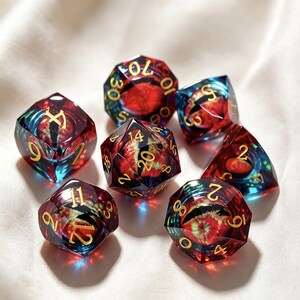 May include: A set of seven polyhedral dice made of translucent resin with a deep red and blue swirling color pattern. Each die features sharp edges and gold-colored engraved numbers. The set includes various shapes including a d20, d12, d10, d8, d6, and d4.