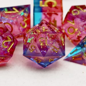 Pode incluir: Um conjunto de dados poliédricos em tons de rosa, roxo e azul com numeração dourada. Os dados são feitos de resina transparente com flocos de ouro no interior. O conjunto inclui uma variedade de formas, incluindo um d20, d12, d10, d8, d6 e d4.