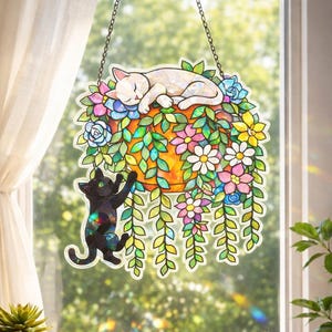 Op de afbeelding: Gebrandschilderde kattenvanger met een slapende witte kat op een bloemstuk en een zwarte kat eronder. Het arrangement heeft kleurrijke bloemen en groene bladeren, opgehangen aan een ketting.