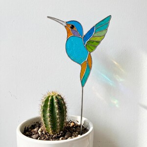 May include: A stained glass hummingbird garden stake with a vibrant design. The bird features a turquoise body, orange accents, and multi-colored wings. The hummingbird is mounted on a metal stake and is placed in a white pot with a cactus.