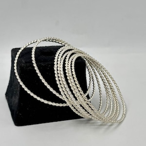 Pode incluir: Uma coleção de pulseiras prateadas com design de corda torcida. As pulseiras são empilhadas em um suporte de exibição de veludo preto sobre um fundo branco. As pulseiras têm um acabamento metálico brilhante.