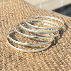 Puede incluir: Seis pulseras plateadas apiladas, de forma redonda y con textura martillada. Las pulseras brillan con un brillo metálico. Se encuentran sobre una superficie marrón texturizada. Un accesorio sencillo y elegante.