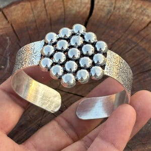 May include: A silver cuff bracelet featuring a central cluster of polished, spherical metal beads. The band of the bracelet displays an intricate, embossed floral pattern on both sides, while the interior surface remains smooth and plain.