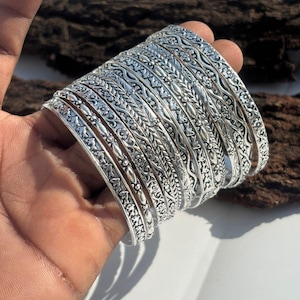 Pode incluir: Um conjunto de pulseiras de prata com intrincados padrões florais e de folhas. As pulseiras são seguradas em uma mão, mostrando os desenhos detalhados e a qualidade reflexiva do metal. As joias são exibidas em um fundo de madeira natural.