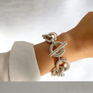 Könnte beinhalten: Ein klobiges Silberkettenarmband mit großen, polierten kreisförmigen Gliedern und einem markanten T-Steg-Verschluss. Das Armband besteht aus dicken, abgerundeten Metallsegmenten, die ein kräftiges, durchgehendes Schlaufendesign bilden.