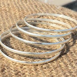 May include: A set of five thin, polished silver-colored bangles stacked together. Each bangle features a smooth, rounded surface with a reflective metallic finish. The bracelets are circular in shape and have a consistent, uniform thickness throughout.