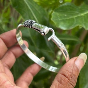Pode incluir: Uma pulseira de metal em tom prateado com uma banda lisa e polida e um fecho de gancho. O mecanismo de fecho é acentuado com um design texturizado de corda torcida no ponto onde as duas extremidades da pulseira se encontram.