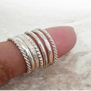 Op de afbeelding: Een verzameling zilveren ringen in verschillende ontwerpen, waaronder gedraaide touwen, gehamerd en gegraveerd. De ringen zijn op elkaar gestapeld en tonen verschillende texturen en stijlen. De sieraden worden op een vinger getoond.
