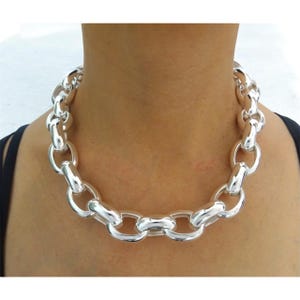 May include: A chunky silver-toned chain necklace featuring large, polished, oval-shaped interlocking links. The necklace sits closely around the neck, showcasing a high-shine metallic finish and a uniform, heavy-duty link design throughout.