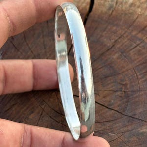 May include: A polished silver-colored bangle bracelet with a smooth, rounded exterior surface and a reflective metallic finish. The bracelet features a simple, continuous circular band design with no additional patterns, engravings, or decorative elements.