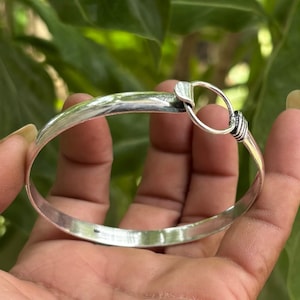 Puede incluir: Un brazalete rígido de tono plateado pulido con una banda curva y lisa que termina en un cierre de anillo circular. El mecanismo de cierre incluye un pequeño detalle de metal texturizado donde el anillo se conecta a la banda principal.