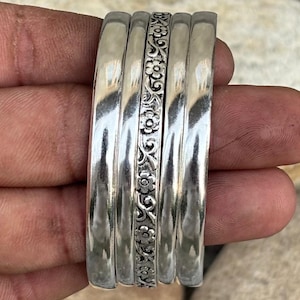 May include: A set of five silver-toned bangles. The outer two bangles on each side have a smooth, polished, reflective surface. The central bangle features a detailed, repeating embossed floral pattern with small flowers and vines.