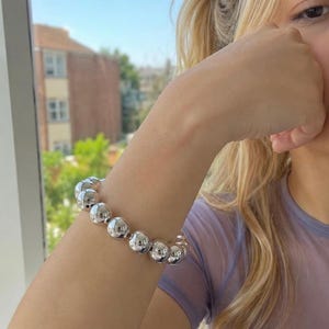 Peut inclure: Un bracelet élastique composé de multiples grosses perles sphériques en métal argenté poli. Les perles sont de taille et de forme uniformes, formant un anneau circulaire continu qui reflète la lumière sur sa surface métallique lisse.