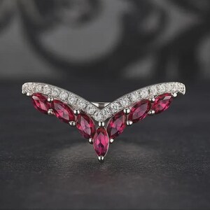 Puede incluir: Un anillo de plata con un diseño en forma de V. El anillo presenta piedras preciosas rojas oscuras con corte marquesa y pequeñas piedras preciosas claras y redondas. El anillo está sobre un fondo oscuro.