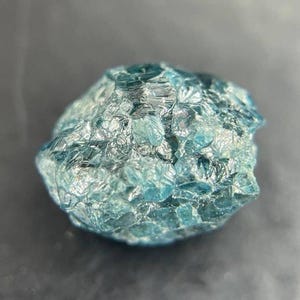 Puede incluir: Una gema en bruto, sin cortar, con una estructura cristalina. La piedra es de color verde azulado claro con facetas reflectantes. La forma general es irregular y texturizada, ligeramente redondeada. La gema parece natural y sin pulir.