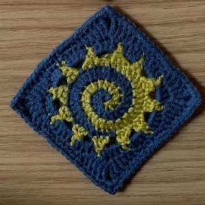 Op de afbeelding: Een vierkant, gehaakt item met een blauwe achtergrond en een gele spiraalvorm in het midden. De spiraal is omgeven door een zonnesterpatroon. Het ligt op een houten oppervlak.