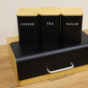 Puede incluir: Una panera negra con asa plateada y tres botes negros con tapas de bambú. Los botes están etiquetados con "COFFEE", "TEA" y "SUGAR" en blanco. La panera y los botes están sobre una superficie de madera.