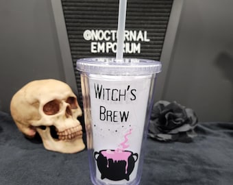 May include: A clear plastic tumbler with a lid and straw, featuring the black text "WITCH'S BREW" above a graphic of a black cauldron bubbling with pink liquid. The design includes small black stars and is positioned upright.
