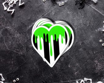 May include: A stack of heart-shaped stickers with a green dripping design over black and white stripes. The stickers are on a dark gray surface with Halloween decorations, including plastic spiders, skeletons, and spiderwebs.