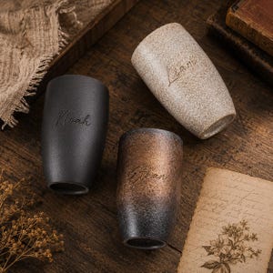 Op de afbeelding: Drie keramische bekers met de namen "Noah", "Liam" en "Ethan". De bekers hebben een getextureerde afwerking in zwart, gespikkeld beige en een brons-naar-zwart verloop. De namen zijn elegant op elke beker gegraveerd.