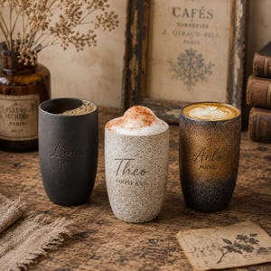 Op de afbeelding: Drie keramische koffiemokken met koffie. De mokken zijn zwart, gespikkeld en bruin met de namen "Luna", "Theo" en "Arlo" erop gedrukt. De mokken zijn gevuld met koffie en latte art. Een vintage-achtige ingelijste print staat op de achtergrond.
