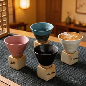 May include: Four ceramic coffee cups in pink, blue, black, and white, each resting on a small, square wooden base. The pink cup is labeled "Billy", the black cup "custom", and the white cup "Kendy". The blue cup has a brown swirl design.