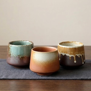 May include: Three ceramic teacups with a rustic design. The cups have a variety of colours, including teal, orange, and brown. The cups are sitting on a grey cloth on a wooden table.