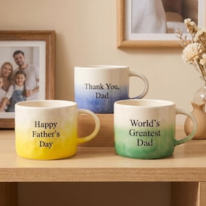 Könnte beinhalten: Drei Keramikbecher mit strukturierter Oberfläche und Farbverlauf. Der gelbe Becher trägt die Aufschrift Happy Father's Day, der blaue Thank You, Dad und der grüne World's Greatest Dad in schwarzer Schrift.