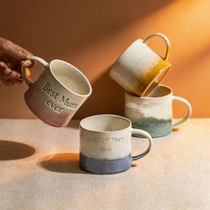 May include: Several ceramic mugs in shades of white, blue, and yellow. One mug, held by a hand, is inscribed with "Best Mum ever." Another mug features the text "CUSTOM TEXT Here." The mugs have a speckled finish.