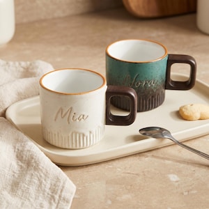 May include: Two ceramic mugs with square handles and ribbed bases. One mug is cream with the name Mia, and the other is teal and brown with the name Nora. Both have white interiors and orange rims, sitting on a rectangular tray with a spoon.