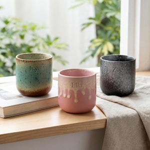May include: Three cylindrical ceramic cups with personalised names engraved on the front. The left cup is teal and brown with a speckled finish and the name Ivy. The centre cup is pink with a cream drip glaze and the name Ella. The right cup is dark grey with a speckled finish and the name Zoe.