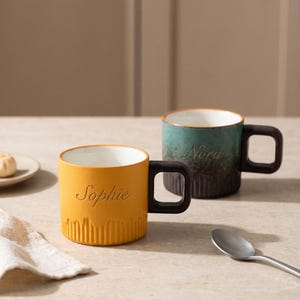May include: Two ceramic mugs with square handles and vertical ribbed bases. The front mug is mustard yellow with the name Sophie engraved in script. The back mug is a teal and dark brown gradient with the name Nora engraved in script.