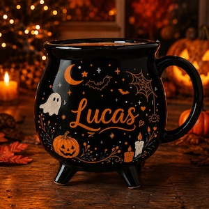 May include: A black ceramic cauldron-shaped mug featuring the name Lucas in orange script. The mug is decorated with orange and white illustrations of a crescent moon, stars, a ghost, a spider web, a jack-o'-lantern, and lit candles.