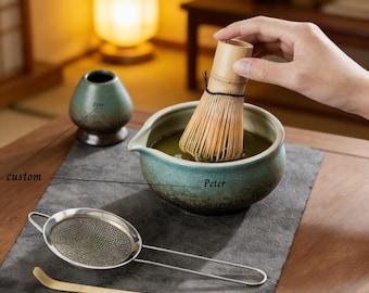 May include: A matcha tea set featuring a bamboo whisk, a ceramic bowl with green tea, a small ceramic tea cup, a metal strainer, and a bamboo scoop. The set is arranged on a dark gray surface, with a warm, inviting background.