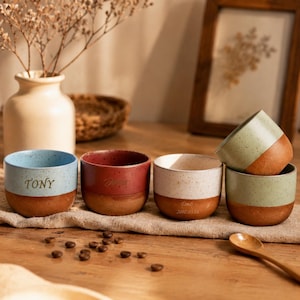 May include: A set of five ceramic cups with a speckled finish and a two-tone design. The cups feature a smooth coloured upper section and a textured terracotta base. Three cups are personalised with the names TONY, Timo, and the date 2002.05.18.