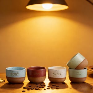 May include: A collection of personalized ceramic coffee mugs with a speckled finish. Each mug has a two-tone design, with a light-colored top and a brown base. The mugs are inscribed with names: Tony, Jess, Susan, and another name. Coffee beans are scattered around the mugs.