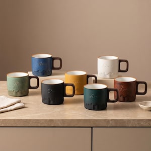May include: A collection of seven ceramic mugs in various colours including blue, green, yellow, black, white, and brown. Each mug features a white interior, a square-shaped handle, and a textured base with vertical ridges and a name engraved.