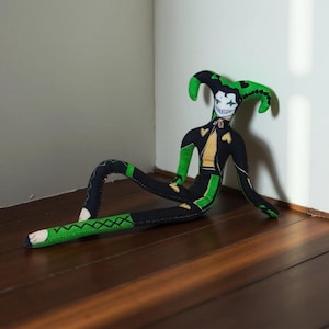 Puede incluir: Muñeca de tela hecha a mano con forma de bufón, con cuerpo negro, detalles verdes y chaleco amarillo. La muñeca bufón tiene una cara blanca con una sonrisa pintada y un gorro de bufón verde. La muñeca está tumbada sobre una superficie de madera.