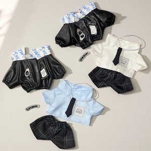 Puede incluir: Se muestran varios conjuntos de muñecas, que incluyen tops y pantalones cortos de cuero sintético negro con cremalleras y esposas, y camisas blancas y azul claro con corbatas negras y pantalones cortos a cuadros. Los conjuntos tienen pequeñas etiquetas y las palabras "DOG BABY" y "CAT BABY".