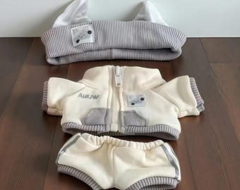 May include: A three-piece doll outfit in cream and gray. The set includes a hooded hat with bear ears, a zip-up jacket with the word "AHUW", and shorts. The jacket and shorts have gray accents. The hat has a gray and white striped pattern.