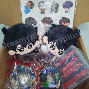 Puede incluir: Una caja de cartón llena de productos de anime. Contiene dos muñecos de peluche con pelo negro, insignias de personajes, llaveros y pegatinas. Las insignias y los llaveros tienen diseños holográficos. La caja está llena de cintas rojas.
