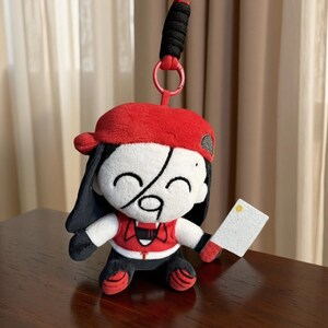 Puede incluir: Una muñeca de peluche con gorra roja, pelo negro y un atuendo rojo y blanco. La muñeca sostiene un pequeño rectángulo blanco y tiene un cordón negro sujeto a un anillo rojo. La cara de la muñeca tiene un diseño único.