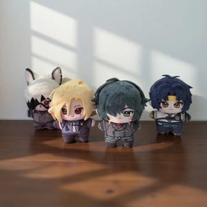 May include: Four plush dolls with anime-style features are displayed on a wooden surface. The dolls have different hair colours and outfits, including a doll with a raccoon-like appearance. The dolls are approximately 15 cm tall.