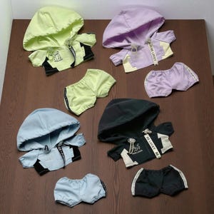 Puede incluir: Conjunto de sudaderas con capucha y pantalones cortos para niños en varios colores pastel, incluyendo verde, morado, azul y negro. Cada conjunto presenta una sudadera con cremallera y pantalones cortos a juego. Las sudaderas tienen una franja blanca con el texto "BE LUCKY".