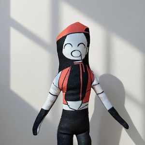 Puede incluir: Muñeca hecha a mano con un diseño estilizado. La muñeca presenta una gorra roja, cabello negro y una cara sonriente. Viste un chaleco negro y naranja, pantalones negros y guantes negros. La muñeca está hecha de tela y tiene una estética simple y caricaturesca.