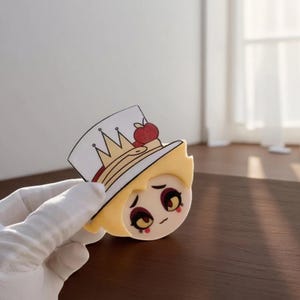 May include: A cartoon-style character head with a white hat featuring a gold crown and a red apple. The character has blonde hair, large eyes, and a sad expression. The image is a close-up of a cut-out figure.