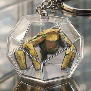 May include: A geometric keychain with a silver-colored ring and chain. The clear, faceted pendant encases a golden-brown, stylized figure with green accents. The keychain is reflected on a shiny surface.