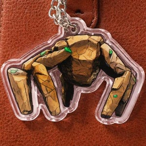 May include: A clear acrylic keychain featuring a brown, stone-like figure with green accents, hanging from a silver chain and ring. The keychain is attached to a brown leather handbag.
